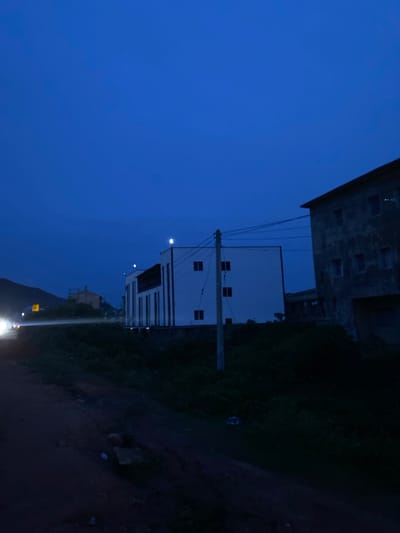 The image depicts a street scene at dusk in Gwong, Nigeria. A dirt road curves through the foreground, bordered by dark vegetation. To the right, a weathered, multi-story building with visible windows stands prominently. In the middle ground, a modern, white building with a dark roof and numerous windows is visible. A utility pole with wires extends from the foreground towards the buildings. In the distance on the left, a vehicle with its headlights on illuminates the road, and a hill can be seen behind it. The sky is a deep, dark blue, indicating late evening or early morning twilight.