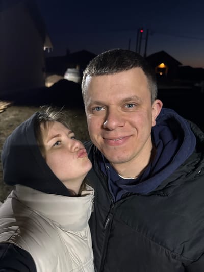 The image shows a selfie of two people, a man and a woman, likely taken in an outdoor setting that appears to be Gornyi (Dvurechenskoe S/P), Russia. The man is wearing a black winter coat over a dark blue hooded sweatshirt. He has short, dark hair and blue eyes, and is smiling at the camera. The woman, positioned slightly below and to the right of the man, is wearing a light beige puffer jacket and a black hooded sweatshirt. She has light-colored hair and is looking upwards with her lips pursed in a kissing expression. The background is dark, suggesting it might be evening or night. Some faint shapes of buildings or structures are visible in the distance, along with what appears to be a streetlight. The ground is dimly lit.