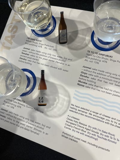 This image is a close-up of a table top with various bottles and glasses of sake, accompanied by printed descriptions of their origin and production methods. The location is in Hamilton, Australia, at coordinates -27.443 latitude and 153.075 longitude. The environment is a brightly lit, white table, and the bottles are labeled with Japanese script alongside descriptions in English. The scene depicts a selection of sakes for tasting, possibly at a restaurant or event.  There are two glasses of sake, one partially full and the other empty. There is also a bottle of sake with a label that reads "I'm Sake Rock, Mifuku Shuzo, Shiga Prefecture" and another bottle labeled "Tsuchida Sake Brewery, Gunma Prefecture". Next to the bottles are printed descriptions of the sake types, including "Junmai," "Ginjo," "Genshu," "Muroka," and "Nama."  The descriptions provide details about the sake's production, including the use of rice, water, koji, and yeast, as well as the addition or absence of brewers alcohol.  The text also mentions the use of ancient yeast starters and refers to specific brewing techniques.  In addition to the sake, the image also includes a text about cheese and cured meat. It reads "We have selected a range of cheese and cured tasting seminar. We encourage you to try a selected a few recommendations with the cheese."  The text lists four cheeses: Ossau Iraty Basque, Wyngaard Chevre Aged Gouda, Creamy French Blue (try with Tsuchida Kimoto), and Comte (try me with everything!). The text also mentions "portions of cured meat, including prosciutto."  This image depicts a tasting event where guests can sample different varieties of sake, paired with cheese and cured meat.