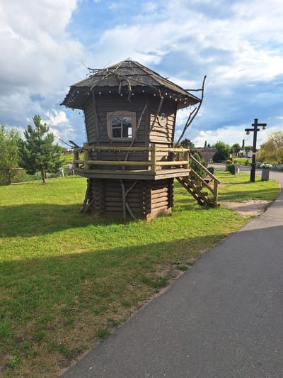 The image depicts a whimsical, rustic wooden structure resembling a small, multi-level hut or birdhouse, situated in an outdoor setting. The hut is elevated on a base constructed from logs and features a wooden deck with a railing, accessible by a wooden staircase. Decorative string lights adorn the railing and the roof's eaves. The roof is thatched and adorned with dried branches. A single window with a lattice design is visible on the front of the hut. The hut stands on a well-maintained green lawn. In the background, various trees, fences, and a lamppost with two light fixtures can be seen. The sky is partly cloudy with a mix of blue sky and dramatic clouds. The scene is located in Малахавецкі сельскі Савет, Belarus, implying it might be a feature in a park, recreational area, or a unique architectural element within a village. There are no people visible in the image, and no actions are taking place. The overall impression is one of a charming, stylized construction in a rural or park-like environment.