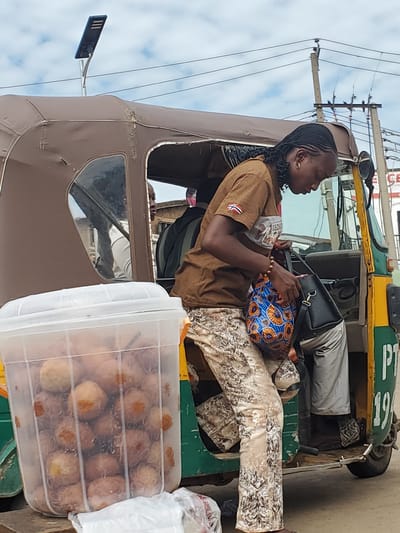 In Gwong, Nigeria, a woman with braided hair is seen disembarking from a brown and green auto-rickshaw. She is wearing a brown t-shirt and patterned trousers, and is holding a black handbag and a colorful patterned tote bag. A large, clear plastic container filled with round, fried food items is positioned in the foreground to the left of the auto-rickshaw. The background shows a street scene with power lines and a solar-powered lamp post.