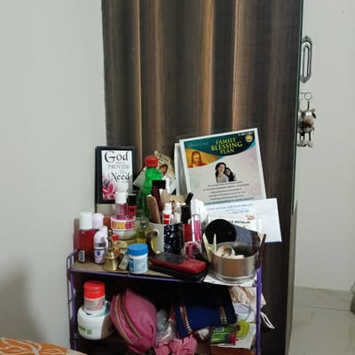 The image shows a cluttered two-tiered shelf against a wall and a door in what appears to be a home in Bengaluru, India. The top shelf is crowded with various items, including cosmetic products, bottles, a small brass bell, and a red mobile phone. A framed picture with the text "God Shall Provide All Your Need" and a "Jesus Calls Family Blessing Plan" leaflet are also present. The bottom shelf contains a pink cosmetic bag, a blue cloth, and other miscellaneous items. The environment suggests a personal space, possibly a bedroom. The door is partially visible on the right, with a door handle and a cartoon keychain hanging from it.