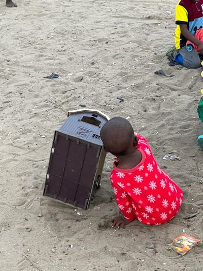 The scene depicts a child in red pajamas with white snowflake patterns kneeling on a sandy surface, likely a beach, next to a brown and grey toy barn. The child appears to be examining the barn. Another individual, partially visible, is wearing a yellow and black sleeve and grey pants. The sandy ground is littered with small debris and remnants of plastic. The location is Badagry, Nigeria.