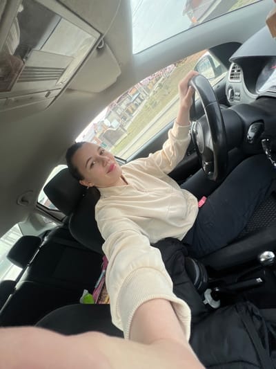A young adult female is depicted taking a selfie from within the driver's seat of a vehicle in the city of Leninavan, Russia. She is smiling and looking directly at the camera, holding a mobile device with her extended right arm. Her left hand rests on the vehicle's black steering wheel. She wears a light beige, ribbed, full-zip sweatshirt and dark pants; a small pink object is visible on her left thigh. The vehicle's interior features black seats, a gray overhead console, and a partially visible dashboard with gauges. Through the windows, a residential street is visible, featuring houses with sloped roofs and sparse vegetation under an overcast sky. A black jacket, possibly headphones, and colorful packages are present on the passenger floor and rear seat respectively.