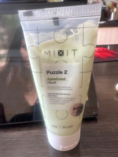 A tube of "MIXXIT Puzzle 2 AquaLand Mask" is positioned on a dark, textured surface. The tube is made of transparent plastic with a puzzle-piece design printed on it, and it contains a pale green cream. Text on the tube includes "Puzzle 2 AquaLand Mask", "ВИТАМИННЫЙ КОМПЛЕКС + ГИАЛУРОНОВАЯ КИСЛОТА", "УВЛАЖНЯЮЩАЯ МАСКА ДЛЯ ЛИЦА", and bullet points in Russian describing its benefits. A circular label indicates "95% NATURAL INGREDIENTS". The bottom of the tube reads "Face | 60 ml e". The background is out of focus, showing reflections on a dark surface, possibly a screen, and hints of a kitchen environment with stainless steel appliances and red objects. The image is captured from a slightly elevated angle. The location is stated to be in the city of Novoaltaysk, Russia.