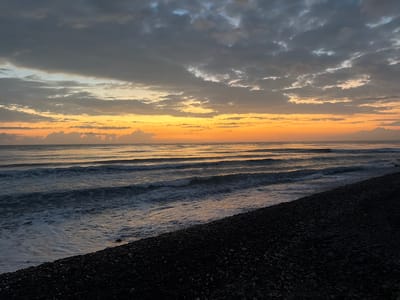 The image shows a natural landscape of a beach during a sunset. The sky is a gradient of colors, transitioning from dark gray clouds at the top to bright orange and yellow hues near the horizon. The sea is visible with gentle waves rolling towards the shore. The foreground consists of a dark, pebbly beach. The overall scene depicts a serene and picturesque coastal environment. The location of Abang, Indonesia, is not directly identifiable from the image itself, as there are no specific landmarks or contextual clues to confirm it.