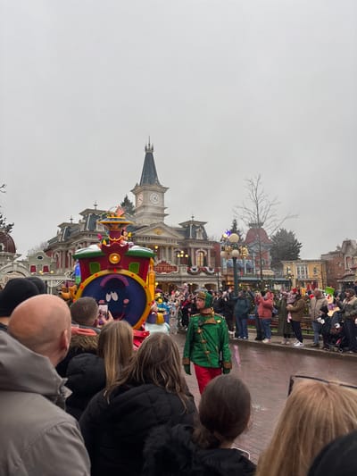 An outdoor scene depicts a parade in progress on Main Street, U.S.A. at Disneyland Park in Chessy, France, under an overcast sky. The ground appears wet, reflecting light.
In the foreground, the backs of numerous spectators are visible, occupying the lower half of the frame. A bald man in a grey jacket is prominent on the far left. Individuals wear various winter garments, including dark jackets and hats. Several people are holding up smartphones, aimed towards the parade route.
In the mid-ground, a colorful, train-themed parade float moves from left to right. The float features a large blue circular face with white eyes and an open mouth, adorned with festive elements such as artificial snow and gift boxes. A figure resembling Mickey Mouse is positioned at the top of the float. Walking beside the float on the right is a male performer dressed in a green, sequined uniform with gold trim, red pants, and a matching green hat, looking towards the right side of the parade route. Beyond the performer, another line of spectators observes the event, some actively recording with phones, while others watch.
The background is dominated by the Main Street City Hall building, characterized by its ornate architecture, including a central clock tower with a dark spire. The building facade features windows, columns, and a visible sign that reads "CITY HALL." Festive garlands and decorations are draped across the buildings lining the street. Leafless trees are scattered between structures.