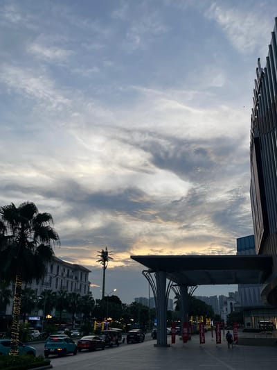 This image captures an outdoor scene in Hanoi, Vietnam, likely during the late afternoon or early evening as suggested by the warm hues in the sky. The sky is filled with dramatic, wispy clouds, some appearing dark and heavy, while others are illuminated by the setting sun, creating a striking contrast.
In the foreground, the edge of a modern building with a distinctive angled, metallic facade is visible at the top. Below this, a paved walkway runs alongside a street. Several red "SALE" banners are displayed prominently on stands along the walkway, indicating a commercial area, possibly a shopping district or a street market.
The scene includes parked vehicles, including cars and motorcycles, suggesting urban traffic. Palm trees are scattered throughout the scene, adding a tropical element to the cityscape. In the background, various buildings of different architectural styles are visible, some appearing to be residential or office blocks. Streetlights and decorative lights are also visible, adding to the ambiance as dusk approaches.
There are a few people visible, including someone standing near the "SALE" signs, possibly a vendor or a shopper. Another person appears to be walking further down the street. The overall impression is a busy urban environment during a visually dynamic time of day, with elements of commerce and tropical flora.