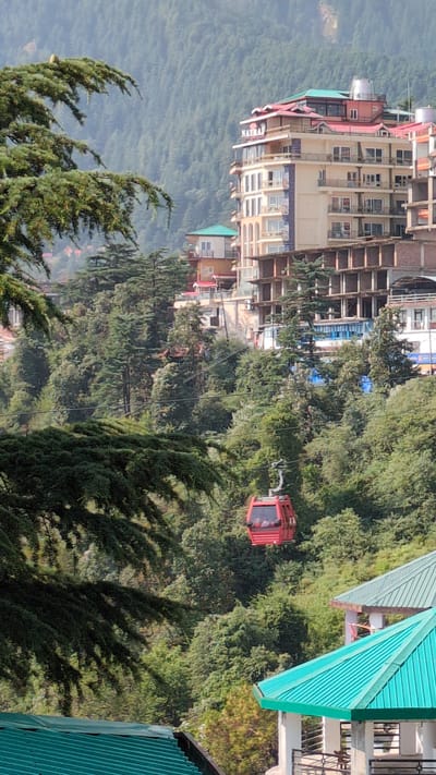 The image depicts a hillside scene in Dharamshala, India. The scene includes a red cable car suspended in the air, against a backdrop of dense trees and buildings. The architecture of the buildings suggests a mountainous region, with structures built along the slope. Some structures bear signage, while others are under construction or appear older. In the foreground, coniferous trees create shadows and texture. In the background, the hills form a scenic mountainous landscape.