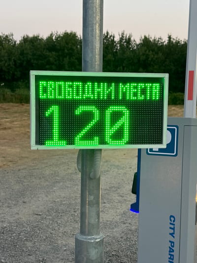 The image shows a bright green LED display mounted on a metal pole. The display shows the Cyrillic text "СВОБОДНИ МЕСТА" above the number "120". To the right of the pole is a gray parking payment machine with "CITY PARK" written vertically in blue letters. The background consists of trees and a pale sky. The ground is covered with gravel. The scene appears to be an outdoor parking area.