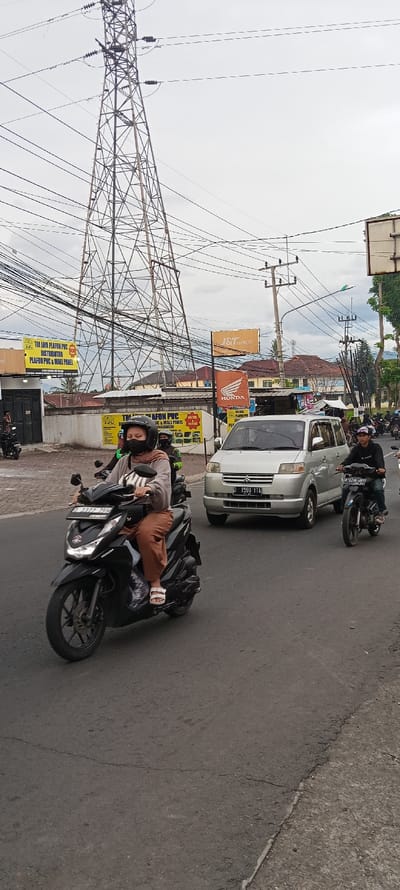 The image shows a street scene in Cangkuang, Indonesia, located at -7.044 latitude and 107.56 longitude. In the foreground, a woman wearing a brown hijab and a black face mask is riding a black motorcycle. The motorcycle is in the left lane of the street. Behind the woman, a white minivan is driving in the right lane. A man on a black motorcycle with a helmet and a green jacket is stopped behind the minivan. In the background, a large metal electrical tower stands tall with numerous power lines spanning across the sky. The road is asphalt and the sidewalk is concrete. The sky is overcast with a few wispy clouds. There are signs for shops and businesses in the background. Several trees and buildings are visible. The woman is riding the motorcycle towards the viewer.