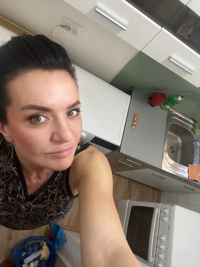 The image displays a rotated indoor scene, approximately 90 degrees counter-clockwise, centered on a woman taking a selfie within a residential kitchen located in Smederevska Palanka Municipality, Serbia.

**Person:**
A woman, appearing to be middle-aged, occupies the left and central portion of the frame. She has dark hair styled away from her face, exposing her forehead and ears. Her skin tone is light, and she wears visible makeup, including eyeshadow, eyeliner, mascara, and blush. Her eyes are brown, and she looks directly at the camera with a subtle expression. She is wearing a dark, sleeveless top featuring a light-colored, intricate abstract or floral pattern. A delicate, gold-colored necklace with small, dark charms is visible around her neck. Her right arm and shoulder are visible in the lower-right quadrant, extending towards the camera, indicating she is holding the device herself.

**Environment and Objects:**
The background reveals a kitchen setting.
*   **Upper-left quadrant:** A white double electrical outlet with a light brown power cable plugged into one socket and extending upwards out of frame. This outlet is affixed to a white surface, likely a wall or upper cabinet side.
*   **Upper-right quadrant:** White overhead kitchen cabinets with horizontal silver bar handles. One cabinet door appears to be a darker grey or black material.
*   **Mid-left:** A white appliance, possibly a washing machine or dishwasher, is partially visible.
*   **Mid-right:** A kitchen counter area featuring:
    *   A dark green wall section, likely tiled or painted, above the counter.
    *   A grey countertop.
    *   A built-in stainless steel sink with a faucet.
    *   Items on the counter near the sink include a red plastic cup, a green bottle (likely dish soap) with a white cap, and a small orange square item (possibly a sponge or cloth).
    *   A brown rectangular package or bar is attached to the side of a grey base cabinet.
*   **Lower-left quadrant:** A blue plastic bag, open at the top, containing assorted refuse, including plastic bottles and other items, rests on the floor.
*   **Lower-middle/right:** The floor is covered with light-colored wooden planks.
*   **Below the woman's right arm:** A white microwave oven with a dark front door and white control knobs is visible.

The lighting appears to be ambient indoor illumination. No external light sources are directly visible.