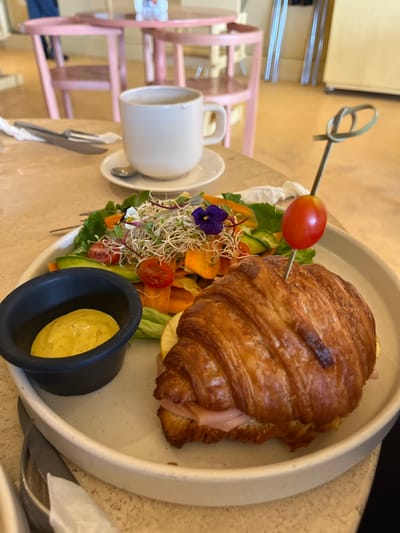 This image depicts a prepared meal set on a table in an indoor dining environment, specifically located in San Andrés Cholula, Mexico. The central subject is a light-colored, circular ceramic plate holding a large, golden-brown croissant sandwich filled with pink ham slices. A thin, dark-colored skewer pierces the croissant, topped with a single red cherry tomato and a green decorative loop. Alongside the croissant is a fresh side salad comprising green leafy lettuce, thinly sliced orange carrots, green cucumber slices, halved red cherry tomatoes, white sprouts, and two small edible flowers, one purple and one with white and yellow petals. A small, matte black ramekin to the left of the salad contains a light yellow, creamy sauce. In the upper left background, a white ceramic mug with a handle rests on a matching saucer, which also holds a silver spoon. To the left of the mug, a silver fork and knife are partially visible, wrapped in a white napkin. The background is blurred, revealing pink wooden chairs and tables, a warm-toned floor, and indistinct metallic structures, indicative of a cafe or restaurant setting. No individuals are visible in the frame.