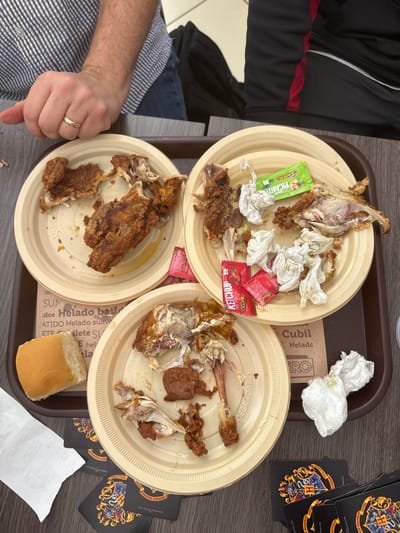Three tan plates stacked on a brown tray contain remnants of a meal, including chicken bones, torn napkins, and condiment packets. Ketchup and a green "PICANTE CAMPERO" packet are visible. A partially eaten bread roll sits on the edge of the tray. A wad of used napkins is near the top. The tray also has text printed on it: "Cubil Helado" and "DEGRADABLE." Playing cards with a crest are scattered around the tray, and a hand with a wedding ring gives a thumbs-down gesture.