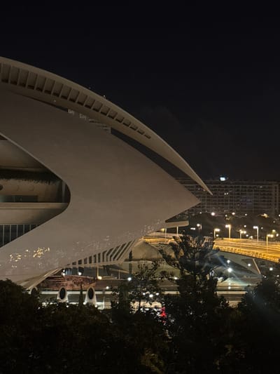 The image, taken at night in Valencia, Spain, captures a section of the striking architecture of the City of Arts and Sciences, specifically the Hemisfèric. The primary subject is the building's distinctive eye-shaped structure, made of concrete and glass, which appears to be partially illuminated. The building's sleek, white, curved form dominates the foreground, with its open underside revealing glimpses of interior elements and an arched entranceway. Greenery, likely a vertical garden or vines, can be seen growing within this arch.
In the background, a tall, modern building with a grid-like facade is visible, its windows lit, suggesting it is an office or residential tower. A elevated roadway, lined with streetlights, curves through the scene, with several poles supporting lights casting a warm glow on the infrastructure. The sky is dark, indicative of nighttime. The presence of palm trees, silhouetted against the lights, adds to the Mediterranean setting. The lighting creates a dramatic, almost ethereal ambiance. No people are visible. The image is slightly tilted.