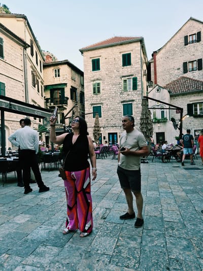 The image depicts a square in Kotor, Montenegro, characterized by its old stone buildings with numerous windows and shutters, and a paved stone ground. In the foreground, a woman is holding up a smartphone to take a picture. She is wearing a black sleeveless top and brightly colored, patterned wide-leg pants, with silver sandals. Beside her, a man stands looking to his left, holding a smartphone in his right hand and a cigarette in his mouth. He is wearing a light-colored t-shirt and dark shorts. In the background, tables and chairs are set up outside a restaurant, with awnings and umbrellas providing shade. Other people are visible seated at tables and walking around the square. The overall atmosphere suggests a warm day in an urban setting.
