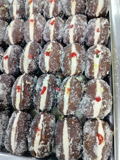 The image shows a close-up view of a tray of Gulab Jamun, a popular Bengali sweet, in Dhaka, Bangladesh. The sweets are arranged in rows and are covered in white sugar crystals. They are dark brown in color with a white filling and are garnished with red, green, and yellow candied fruit pieces. Each Gulab Jamun appears to be approximately 3 cm in diameter and 5 cm in length. They are displayed on a silver tray.