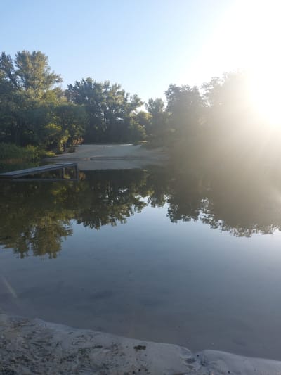 This is a photograph of a tranquil lake scene on a clear, sunny day. The water is calm and reflects the surrounding trees and a bright sunburst in the upper right corner. In the foreground, the sandy shore is visible, with a wooden pier extending into the water on the left side of the image. The background features a line of lush green trees. The scene is devoid of any people or visible activity, suggesting a peaceful, natural setting. It is plausible that this image depicts a location within the city of Kamianske, Ukraine, given the natural elements and clear sky often associated with the region's recreational areas.