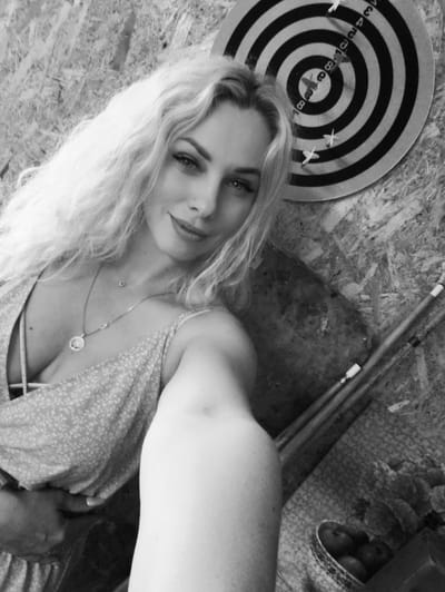 A grayscale selfie captures a young adult female with long, wavy, light-colored hair, a subtle smile, and a nose piercing, looking directly at the camera. She wears a low-cut, light-patterned sleeveless top over a visible undergarment, accessorized with two necklaces, one featuring a circular pendant. Her right arm is extended, holding the camera for the selfie. The background wall is constructed from textured wood, consistent with oriented strand board (OSB). Mounted on the upper right section of the wall is a dartboard with concentric black and white rings and numerical markings, containing several white-fletched darts. Beneath the dartboard, several elongated cylindrical objects, resembling pool cues or painting brushes, lean against the wall. A small bowl or basket containing several round objects, possibly fruits, is partially visible in the lower right foreground. The scene is located in Терновский округ, Ukraine.