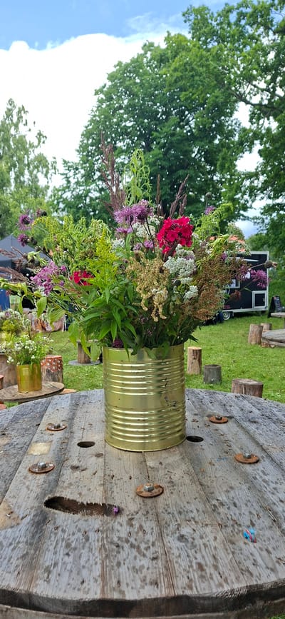 A close-up of a large, gold-colored metal can filled with a vibrant bouquet of wildflowers stands on a weathered, wooden, spool-shaped table. The table has visible bolts and holes, indicating it's likely outdoors. In the background, a grassy area is dotted with tree stumps and other wooden spools, suggesting a park or outdoor event space. Further back, a black food truck with a white trailer is visible, and beyond that, lush green trees under a cloudy sky. The overall setting appears to be a casual outdoor gathering.