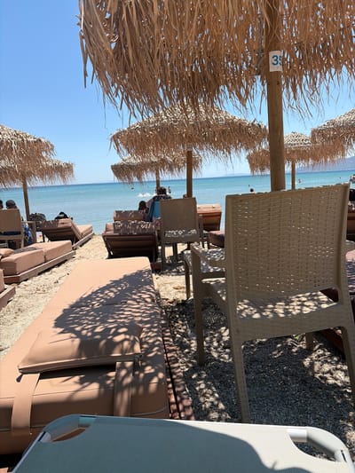 The image depicts a sunny day at a beachside resort, likely in Artemida, Greece. The scene is characterized by rows of sun loungers and straw umbrellas, creating a relaxed vacation atmosphere. The foreground is dominated by a close-up view of straw umbrella material and a portion of a light-colored plastic chair. Behind the chair, several brown sun loungers are visible, some with cushions. The ground is covered with small pebbles.
In the mid-ground, the turquoise waters of the sea stretch out under a clear blue sky. Further back, people can be seen on the beach and in the water, enjoying the sunny weather. The presence of multiple umbrellas and loungers suggests a public or commercial beach. The time of day appears to be late morning or early afternoon, given the bright sunlight and the angle of the shadows cast by the umbrellas and trees.
A small white tag with the number "35" in blue is visible on a wooden support of one of the straw umbrellas, likely indicating a numbered spot or amenity. The overall mood conveyed is one of leisure and relaxation, typical of a beach vacation. The location cues, such as the clear blue sea and the style of the beach setup, are consistent with a Mediterranean setting.
