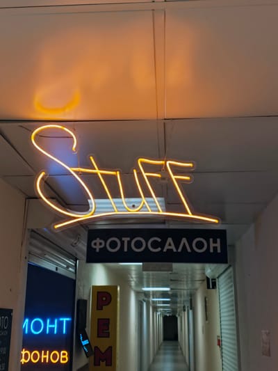The image depicts a long, dimly lit hallway in what appears to be a commercial building. A bright yellow neon sign reading "Stuff" is prominently displayed overhead. Below this, a dark sign with white Cyrillic lettering states "ФОТОСАЛОН" (Photo Salon). To the left, a closed storefront has blue neon lettering that partially reads "МОНТ" (likely part of a word like "Ремонт" - repair). Further down the hallway, on the right, there are closed metal shutters and on the left, more doors and signs are visible, suggesting a row of shops or offices. A yellow sign with red Cyrillic letters "РЕМ" (likely part of "Ремонт" - repair) is visible on the right side of the hallway. The ceiling has fluorescent lighting fixtures. The overall impression is of a slightly dated commercial corridor. The location is specified as Svietlahorsk, Belarus.