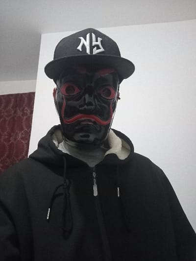 A person is wearing a black and red mask, a black baseball cap with white embroidery, and a black hooded jacket. The mask is dark with red details around the eyes and mouth. The jacket is zipped up and the hood is pulled up, revealing a light-colored lining. The person's face is obscured by the mask. A section of wall is visible to the side, and the edge of a bed or furniture with a dark red and white patterned cover can be seen.