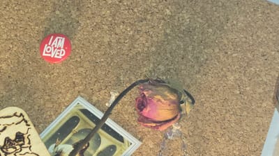 The image shows a close-up of a corkboard. A dried rose with a stem and a necklace sits at the upper left. A postcard with an image of people is at the upper right, next to a wooden decoration. A red button with the phrase "I AM LOVED" is at the lower right. The image does not provide enough information to identify a specific location in Lompoc, United States.