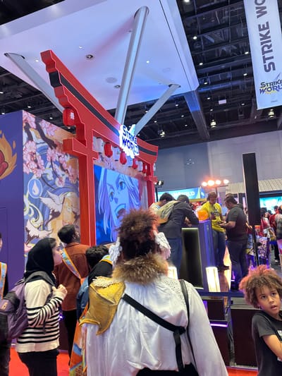 The image depicts an indoor event, possibly a convention or exhibition, centered around a "Strike World" display with Japanese-inspired decorations. The display includes a large red torii gate structure adorned with lanterns and artwork featuring anime-style characters, cherry blossoms, and cranes. Several people are present, including attendees wearing costumes. The location of the scene is NOT possible to discern to be in Mumbai, India, based on the provided image.