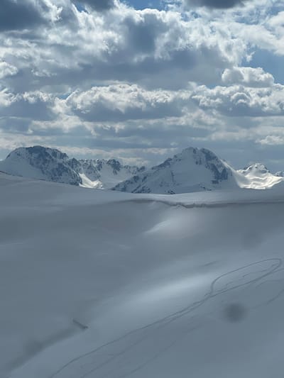 The image displays an expansive, snow-covered alpine environment located in Katun Bačko brdo, Montenegro. The foreground and midground consist of undulating snowfields, presenting a spectrum of white and grey tones due to varied light and shadow. Multiple sets of ski or snowboard tracks are clearly visible on the snow surface in the lower right quadrant, curving across the terrain. The background features a range of rugged, snow-capped mountain peaks with discernible rock formations, extending across the horizon. The sky occupies the upper half of the frame, densely filled with cumulus clouds, interspersed with areas of brighter cloud and patches of blue sky, casting variable illumination across the landscape. No human figures or man-made structures are present, apart from the aforementioned snow tracks.