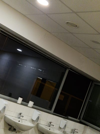 The image depicts a dimly lit public restroom interior, likely at night, as indicated by the dark windows reflecting external lights. The scene features several white ceramic sinks with chrome faucets lined up along a tiled wall. Above the sinks, there are white dispensers, likely for soap or paper towels, and two rolls of toilet paper are precariously placed on the ledge above. The ceiling is a white, textured material with recessed lighting fixtures and what appear to be air vents. A large window runs along the upper portion of the wall, revealing a dark exterior with some faint interior lights visible through it. The overall impression is one of a functional but somewhat neglected public facility. The location is identified as Bahovitsa, Bulgaria.