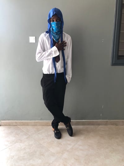 A single individual stands posing indoors against a plain light gray wall in the city of Gbawe, Ghana. The person, appearing male with dark skin, wears a blue durag covering the head and a blue patterned bandana obscuring the lower face, revealing only the eyes. Attire consists of a white long-sleeved shirt, untucked, with a dark patterned tie partially visible beneath the collar. Black trousers are worn, and black backless loafers with gold-toned buckle details are on the feet. The individual's left hand is placed over the chest, while the right hand is inserted into a trouser pocket. The legs are crossed at the ankles, with the right leg in front. The environment features a white square light switch or outlet cover on the left side of the wall and a fragment of a dark-framed window or opening in the upper right portion of the background. The floor is composed of light-colored, possibly beige or cream, square tiles laid with visible grout lines.