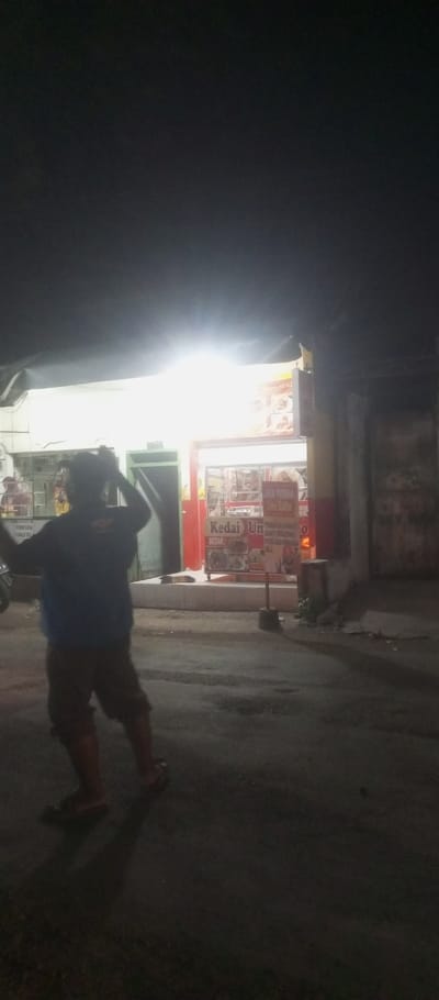 The image shows a man walking away from a storefront in Kecamatan Waru, Indonesia, located at coordinates -7.35 latitude and 112.764 longitude. The man is wearing dark pants and a blue shirt, and is carrying an object in his right hand. The storefront has a red and white sign with the words "Kedai Un" in red letters on a white background. The sign is illuminated by a bright overhead light. The building has a white facade and a few windows. The street in front of the store is paved and dark. The man is walking away from the camera, and his shadow is visible on the ground. The night is clear and the sky is dark.