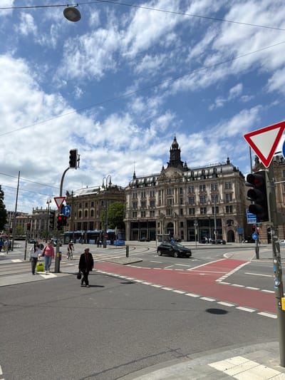 The image depicts a street scene in Munich, Germany, likely near Marienplatz or a similar central urban area, characterized by grand architectural buildings and a busy intersection. The sky is partly cloudy, with scattered white clouds against a clear blue backdrop.
In the foreground, a wide asphalt road features a prominent red-painted pedestrian crossing area with white dashed lines marking its edges. A black car is turning at the intersection. Several pedestrians are visible: two young women with a yellow suitcase walk across the road, and an older person in a dark coat walks towards the viewer. A cyclist on a bicycle is also present in the middle ground.
The background is dominated by impressive historical buildings with ornate facades, classical architecture, and multiple stories. One building, in particular, stands out with a prominent tower and a domed roof. Traffic lights are visible at the intersection, with one red light illuminated, suggesting traffic is stopped. Signs include a yield sign (inverted white triangle with a red border) and directional signs indicating "Passau," "Nürnberg," and "München." A blue tram is stopped at a designated area, and overhead wires for the tram system are visible.
The overall impression is of a vibrant, well-maintained urban environment in a historic European city.