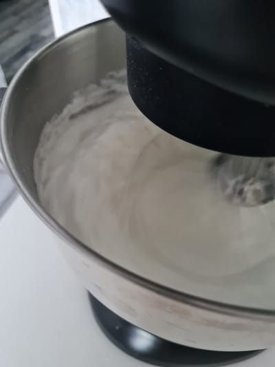A close-up shot shows a stand mixer bowl filled with a fluffy, white substance, likely whipped cream or meringue, in motion. A black attachment, part of the mixer, is visible at the top left, partially obscuring the bowl. The stainless steel bowl shows some smudges and slight discoloration, indicating use. The background is a plain white surface, suggesting an indoor kitchen setting. The lighting is bright, and there are no specific cues for time of day or weather. The image is focused on the texture and movement of the whipped substance. No people are visible, and there is no visible text.