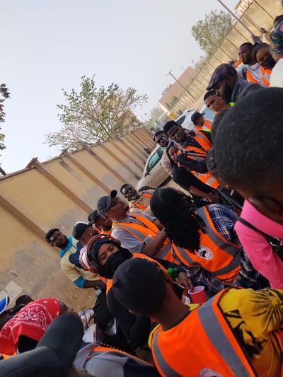 The image depicts a group of approximately 15-20 individuals gathered outdoors in Sabon Gari East, Nigeria. The scene is illuminated by natural daylight, with a pale, overcast sky visible above.

In the background on the left, a light-colored, possibly beige, concrete wall with a patterned top edge extends into the distance. Behind this wall, sparse green foliage from trees is visible. Further in the mid-ground on the right, a light-colored sedan is partially visible, parked or moving, alongside distant structures and a utility pole.

The foreground and mid-ground are populated by the group, consisting primarily of young adults. Many individuals are wearing bright orange safety vests over various casual clothing items, including t-shirts, plaid shirts, and long-sleeved tops. Some vests display a white circular logo on the back. Headwear is common, including black baseball caps, a plaid flat cap, and a red bandana worn around the neck or head. At least one female subject wears a black face mask and headphones or earmuffs. Another individual holds a red plastic cup. Several individuals are engaged in conversation or looking in various directions, some directly towards the camera's perspective. The overall posture of the group suggests a casual gathering.