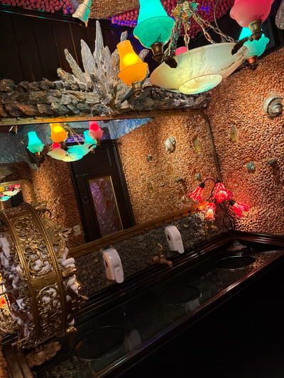 This image depicts the interior of a highly decorated restroom at Gero Restaurant in Ipanema, Rio de Janeiro, Brazil. The walls are textured with a mosaic of numerous small, light brown pebble-like stones, into which various marine shells, including at least one spiral conch, and polished, translucent stone fragments are embedded.
Above a dark, polished counter, a large rectangular mirror is mounted, reflecting the upper part of the room. The reflection shows a dark wooden door with an ornate, possibly stained-glass panel featuring purple patterns. Above the mirror, rock formations resembling coral and geological spikes protrude from the wall. Several chandeliers with multiple arms hold colored glass lampshades in hues of yellow, green, pink, and light blue. A large, flat, shell-shaped light fixture is also visible among these.
The dark counter features multiple circular sink basins, at least two of which are visible. White, wall-mounted soap dispensers are positioned above the counter. Ornate, golden-toned faucets or water spouts are present. On the far left, an elaborate golden-brass towel dispenser with intricate scrollwork holds white rolled towels. On the right side of the counter, a multi-branched lamp with five red, flower-shaped glass shades provides localized illumination. The overall environment is dimly lit, emphasizing the intricate and themed decor. No people are visible.