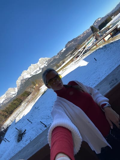 An upward-angled selfie captures a female subject on a balcony overlooking a snow-covered landscape in Going am Wilden Kaiser, Austria, under a clear blue sky. The subject, positioned in the mid-right, has light hair styled in a braid over her left shoulder, wears yellow-tinted sports sunglasses, a red long-sleeved turtleneck, and an open white knitted cardigan. A silver-toned watch is visible on her left wrist. Her right arm extends towards the camera from behind a dark wooden balcony railing in the foreground. The midground reveals a snow-dusted field sloping away, featuring sparse fences, patches of visible grass, and several modern buildings with dark roofs and light facades nestled into the hillside, surrounded by scattered trees. The background is dominated by the majestic, snow-capped peaks of the Wilder Kaiser mountain range under bright sunlight, with prominent rocky faces partially obscured by snow. Shadows are visible on the snow and buildings, indicating a sunny day.