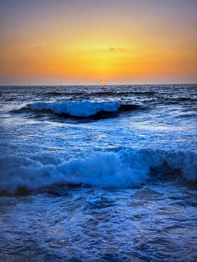 The image depicts a scene where a body of water, potentially the ocean, meets the shore. The left side of the image showcases a soft, orange glow, suggestive of either sunrise or sunset. The water is turbulent with visible waves crashing and churning. No people, objects, actions, or interactions are clearly identifiable in the image. There is no evidence in the image to place it in Bat Yam, Israel.