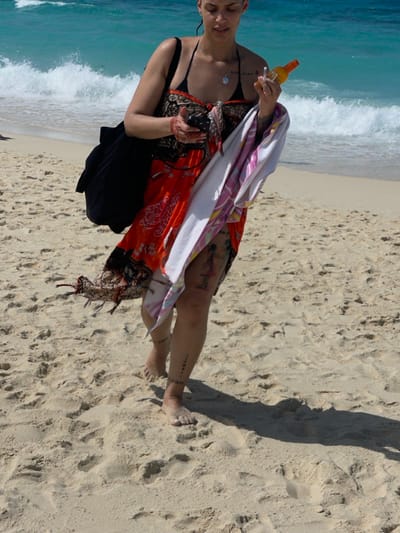 A solitary individual is observed walking barefoot on a sandy beach in the city of Zanzibar, Tanzania. The subject, an adult female with visible tattoos on her arms and legs, wears a black bikini top beneath an orange and black patterned sarong. A black shoulder bag is carried on her left shoulder. In her left hand, she holds a dark-colored smartphone. Her right hand grasps an orange bottle, likely sunscreen, and a small white object, while a white and multi-colored striped towel is draped over her right forearm. The individual casts a long shadow extending to her right on the sand. The environment consists of light-colored sand in the foreground and midground, transitioning to turquoise ocean water with white breaking waves in the background.