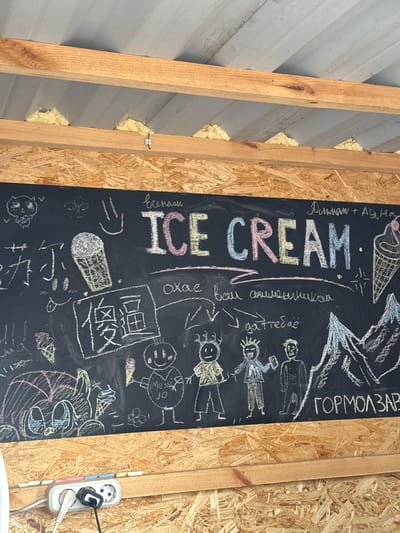 The image is a close-up, slightly angled shot of a blackboard with chalk drawings and text. The blackboard is mounted on a wooden wall, and above it, exposed wooden beams and insulation are visible, suggesting an outdoor or unfinished interior space. The blackboard itself is covered with various drawings, including ice cream cones, cartoon characters, and what appear to be mountains. The main text on the blackboard reads "ICE CREAM" in large, colorful letters. Below "ICE CREAM" is some smaller, handwritten text in what appears to be Cyrillic script. To the right of the "ICE CREAM" text, there is a drawing of an ice cream cone, and further to the right, a mountain range is depicted. The bottom section of the image shows a power strip with two plugs inserted, mounted on a wooden surface. There are also some chalk sticks in various colors lying on the wooden surface in front of the power strip. The overall impression is of a casual, possibly temporary setup for an ice cream vendor. The location is Kokshetau, Kazakhstan.