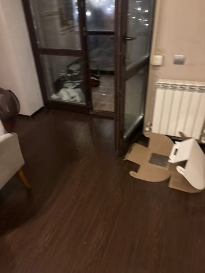 The image depicts an interior scene, likely a room in a residential building. The floor is made of dark wood laminate. To the left, a portion of a light-colored armchair is visible. Dominating the center and right of the image is a dark wooden door with glass panels. Through the glass, a dimly lit interior space, possibly a balcony or another room, is visible, containing some folded white fabric and dark items.
In the foreground, on the right side of the door, are several pieces of flattened cardboard boxes and a white packaging material, suggesting recent unpacking or disposal. A white radiator is mounted on the wall above these boxes. To the right of the door, on the wall, there are two light switches or electrical outlets. The overall lighting is dim and artificial. The scene appears to be indoors, possibly in a living area or hallway, in a city like Orekhovka, Russia, though no specific landmarks confirm this location. There are no people visible in the image.