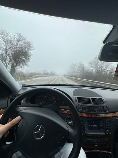 The image presents a forward view from the interior of a Mercedes-Benz vehicle, observing a fog-shrouded road in Palamartsa, Bulgaria. A person's right hand is visible gripping the left side of the black, three-spoke steering wheel, with the thumb positioned on the multi-function controls. The dashboard features a combination of black material and wood grain trim, displaying an instrument cluster and a central console with air vents, climate controls, and a radio interface indicating "87.5 MHz." A circular phone mount is attached to one of the central air vents. Outside the vehicle, a two-lane asphalt road extends into dense fog, which significantly diminishes visibility. Bare trees and low vegetation line both edges of the road, with a metal guardrail present along the right side. The sky is uniformly grey due to the heavy atmospheric mist.