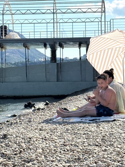 The image depicts a sunny day at a pebble beach in Ut'os, Ukraine. Two individuals are visible: a young boy in the foreground, wearing blue patterned swim trunks, and an adult in the background, partially obscured by an umbrella. The boy is sitting on a towel, looking towards the camera with his hand near his mouth. The adult is seated behind him. They are situated near the water's edge, with the sea visible on the left. A striped umbrella provides shade. In the background, a large concrete structure, likely a pier or embankment, runs parallel to the coastline. Beyond the structure, a hilly landscape with some buildings can be seen under a clear blue sky. The beach is covered in smooth, grey pebbles.