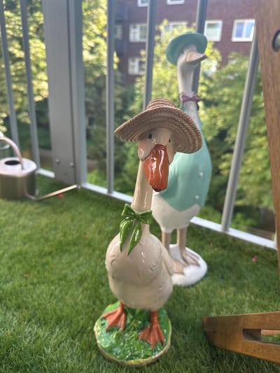 This media file shows two ceramic goose figurines on a patch of artificial grass. The goose in the foreground is wearing a straw hat and a green bow around its neck. Its beak is orange and its eyes are black dots. It stands on a green base with its webbed feet visible. Another goose figurine is partially visible behind it, wearing a blue hat and a purple ribbon. The background is blurred, but appears to be an outdoor setting with trees and buildings. The lighting suggests it is daytime, possibly late afternoon due to the warm tones. The presence of artificial grass and the style of the figurines suggest a garden or patio setting. The context of Hamburg, Germany, is provided as additional information. There is no visible text in the image.