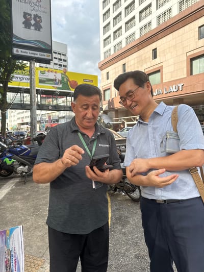 Two men are standing on a sidewalk in Kuala Lumpur, Malaysia, with a building and a billboard visible in the background. The man on the left, wearing a gray polo shirt with a green lanyard, is holding a smartphone and appears to be showing something on the screen to the man on the right. The man on the right, wearing a light blue collared shirt and dark trousers, is looking at the phone and smiling. He has a crossbody bag with a "Kickers" logo. Behind them, a billboard displays an advertisement with cartoon bears and text that reads "Visit Malaysia 2020 Truly Asia". The building in the background has "JA LAUT" written on it. There are motorcycles parked on the sidewalk to the left of the men. The environment suggests an urban setting with mixed lighting, possibly overcast.