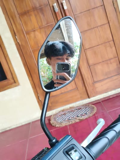 The image shows a reflection of a person taking a selfie in a motorcycle mirror. The person is holding a smartphone, and only part of their face is visible. The background includes a wooden door, a window on the left, and a small rug on a red tiled floor. A glimpse of green foliage can be seen through the mirror, likely outside. The motorcycle's handlebar, including the turn signal switch, is in the foreground. Without more information, it's not possible to specifically confirm or deny its location in Pacet, Indonesia.