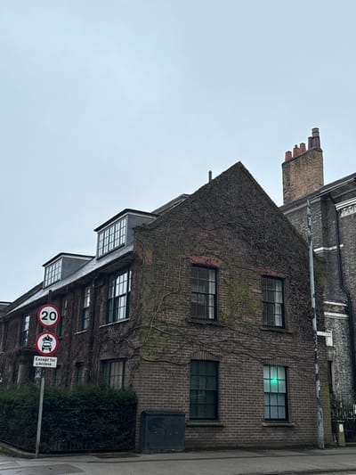 A brick building covered in ivy with several windows. The top window reflects a bright green light. A chimney sits above the building to the left. A street sign is visible at the bottom, displaying a 20 mph speed limit and an exception for access. The sky is overcast.
