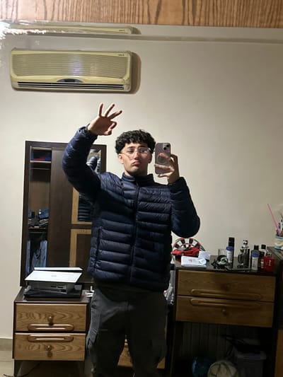 A young man with curly hair and glasses takes a mirror selfie in a room. He wears a dark blue puffer jacket and grey pants, making an "OK" gesture. Behind him is a wooden dresser with perfume bottles, a dartboard, and other objects on top. To the left of the mirror is an air conditioning unit.