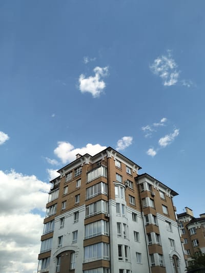 A low-angle shot depicts a multi-story apartment building in Krykhivtsi, Ukraine, against a blue sky dotted with scattered white clouds. The building's facade is characterized by a two-tone color scheme, with the lower stories painted white and the upper stories featuring a brown brick or paneling. Numerous windows punctuate the building's exterior. A second, similar building is partially visible on the right. Overhead, thin power lines stretch across the sky.